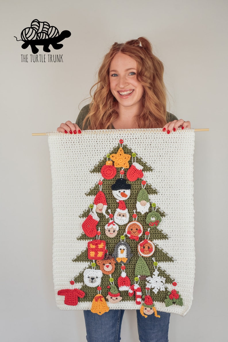 Puede incluir: Un &aacute;rbol de Navidad de crochet blanco y verde con 24 adornos de crochet diferentes colgando de &eacute;l. Los adornos incluyen un mu&ntilde;eco de nieve, Pap&aacute; Noel, un ping&uuml;ino, un hombre de jengibre, una campana, una media, un bast&oacute;n de caramelo y m&aacute;s. El &aacute;rbol est&aacute; colgado de una varilla de madera.