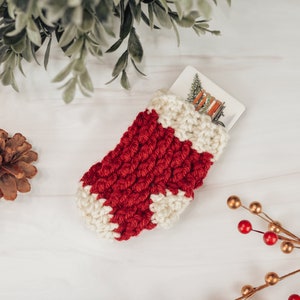 May include: A red and white crocheted stocking with a white card inside. The stocking is on a white wooden surface with pine cones and a sprig of red berries with gold stems.