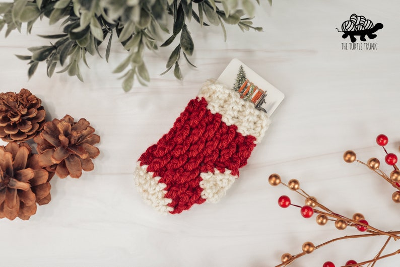May include: A red and white crocheted stocking with a white card inside. The stocking is on a white wooden surface with pine cones and a sprig of red berries with gold stems.