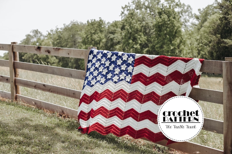 May include: A crocheted American flag blanket draped over a wooden fence. The blanket is red, white, and blue with a chevron pattern and stars. The text "crochet pattern" and "The Turtle Trunk" are visible in a circle on the blanket.