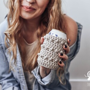 May include: A close-up of a person holding a beige crocheted can cozy. The cozy is made with a textured stitch and has a simple design.