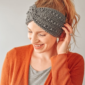 May include: A woman wearing a gray crocheted headband with a twisted design. She is smiling and has her hair pulled back in a ponytail. She is wearing a rust colored cardigan over a gray shirt.