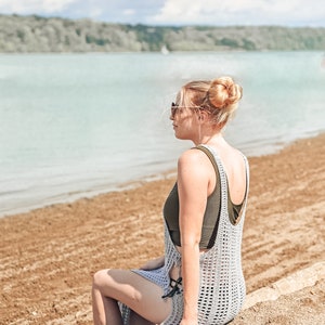 May include: A woman wearing a light grey crochet cover-up with a dark green tank top underneath. She is sitting on a sandy beach with a body of water in the background.