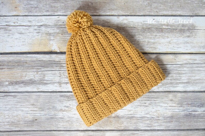 May include: A mustard yellow crocheted beanie with a fluffy pom pom on top. The beanie is lying on a light wood background.
