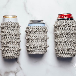 May include: Three crocheted can coolers in a light gray color. The coolers are made with a textured stitch that creates a bumpy surface. The coolers are shown on a white marble surface.