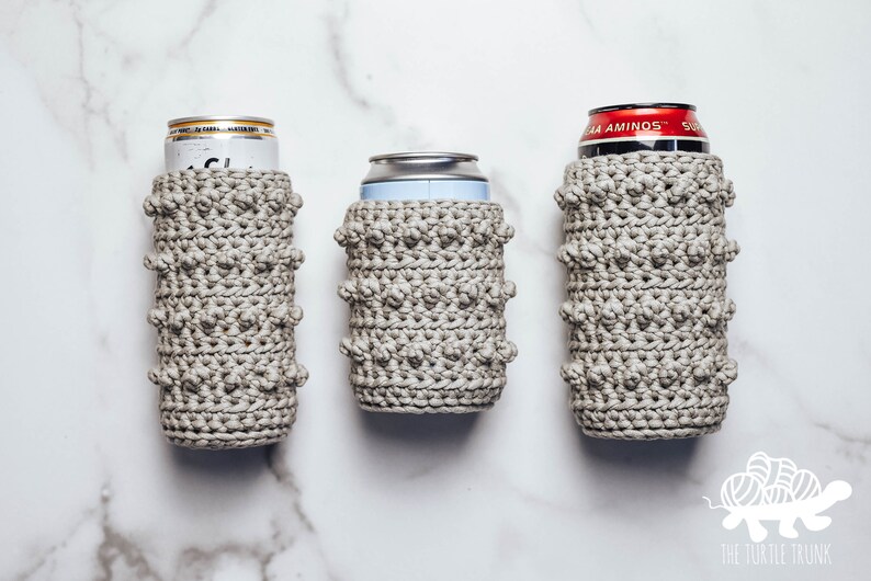 May include: Three crocheted can coolers in a light gray color. The coolers are made with a textured stitch that creates a bumpy surface. The coolers are shown on a white marble surface.