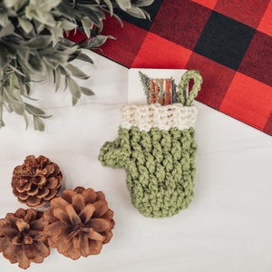 May include: A green crocheted mitten with a white cuff, holding a gift card with a winter scene. The mitten is sitting on a white surface with pine cones and a red and black buffalo plaid fabric.
