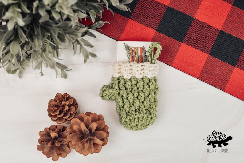 May include: A green crocheted mitten with a white cuff, holding a gift card with a winter scene. The mitten is sitting on a white surface with pine cones and a red and black buffalo plaid fabric.
