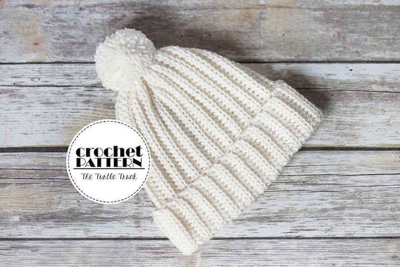May include: White crocheted beanie with a pom pom on top. The beanie is laying on a wooden surface. The text "crochet pattern" and "The Turtle Trunk" is visible on a white circle.