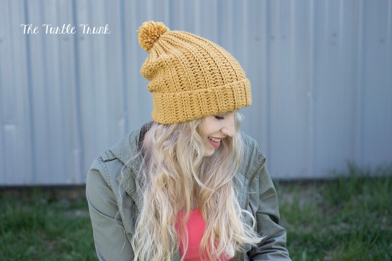 May include: A mustard yellow crochet beanie with a fluffy pom pom on top. The beanie is slouchy and has a ribbed texture.