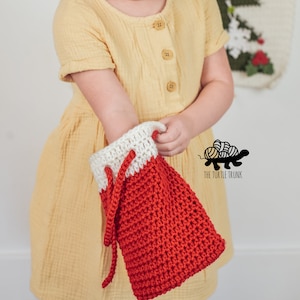 Puede incluir: Una media de Navidad de crochet roja y blanca con cord&oacute;n de ajuste. La media la est&aacute; sujetando una persona que lleva un vestido amarillo.