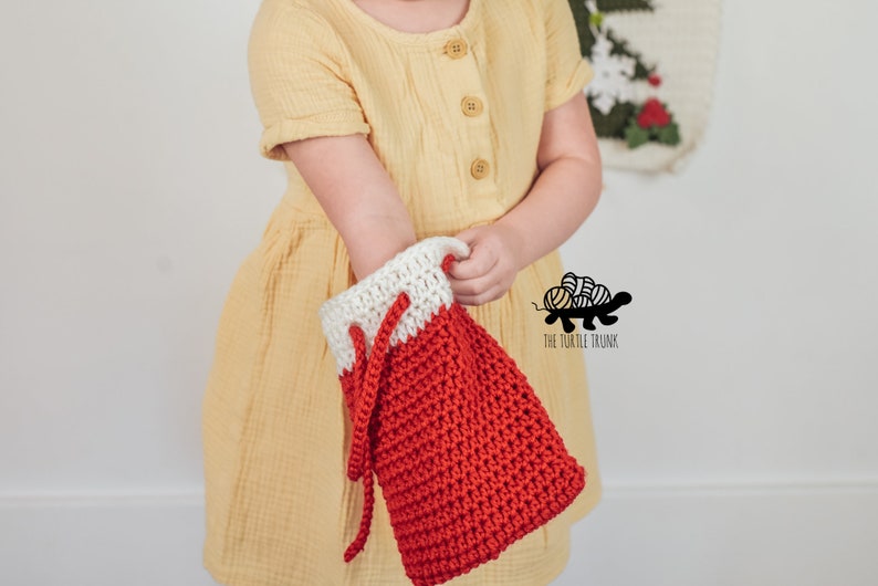 Puede incluir: Una media de Navidad de crochet roja y blanca con cord&oacute;n de ajuste. La media la est&aacute; sujetando una persona que lleva un vestido amarillo.