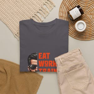 May include: A dark gray t-shirt featuring a graphic of a person's face wearing sunglasses on the left side and the orange text EAT WORK TRAIN stacked vertically on the right side. The shirt has a crew neckline and short sleeves.
