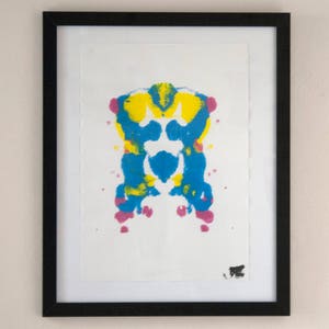 Può includere: Stampa d'arte astratta con un design simmetrico in blu, giallo e rosa. La stampa è incorniciata in una cornice nera.