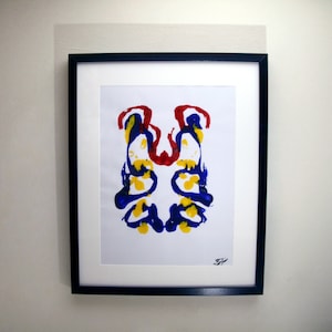 Könnte beinhalten: Gerahmter abstrakter Kunstdruck. Das Kunstwerk zeigt ein symmetrisches Tintenklecksmuster mit roten, gelben und blauen Farbspritzern auf weißem Hintergrund, in einem schwarzen Rahmen.