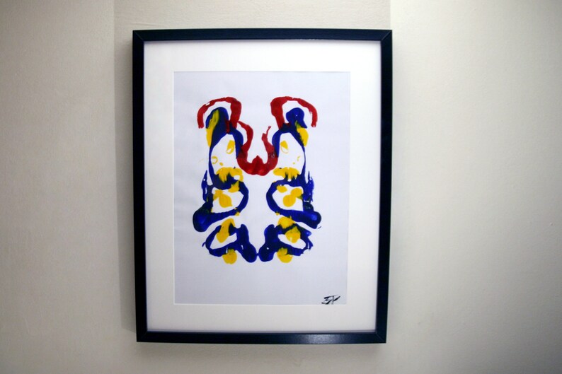 Op de afbeelding: Ingelijste abstracte kunstafdruk. Het kunstwerk toont een symmetrisch ontwerp met blauwe, gele en rode verfspatten op een witte achtergrond. De afdruk is gemonteerd met een witte passe-partout.