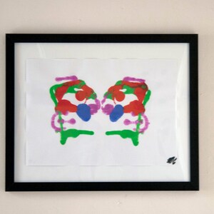 Può includere: Un dipinto astratto incorniciato con un design simmetrico. Il dipinto presenta uno schema di colori rosso, verde, blu e rosa brillanti.