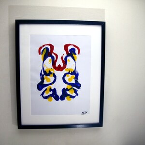 Op de afbeelding: Een ingelijste abstracte inktvlekkenkunstdruk. Het kunstwerk heeft een symmetrisch ontwerp met blauwe, gele en rode inktvlekken op een witte achtergrond. De lijst is zwart en het kunstwerk hangt aan een witte muur.