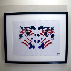 Puede incluir: Una pintura abstracta enmarcada con un diseño simétrico en rojo, azul y blanco. La pintura está enmarcada en negro y tiene un passe-partout blanco.