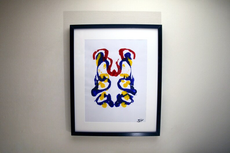 Op de afbeelding: Abstracte kunstafdruk in een zwarte lijst. Het kunstwerk toont een symmetrisch inktvlekontwerp met rode, gele en blauwe verfspatten op een witte achtergrond. De afdruk is gemonteerd op een witte passe-partout.