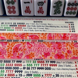 Puede incluir: Un soporte transparente de azulejos de Mahjong de acrílico con un estampado floral y de langosta rosa y naranja. El nombre "Suzanne" está escrito en cursiva. El soporte se encuentra sobre una superficie blanca con fichas de Mahjong e instrucciones del juego.