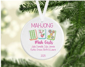 Op de afbeelding: Een rond wit keramieken ornament dat aan een wit lint hangt. Het bevat de tekst MAHJONG en Mah Girls in het roze, met drie mahjongtegels met bamboe-, bloem- en cirkelontwerpen. Daaronder staan de namen Julia, Danielle, Isla, Jennie, Katie, Grace, Beth & Laura en het jaartal 2025.