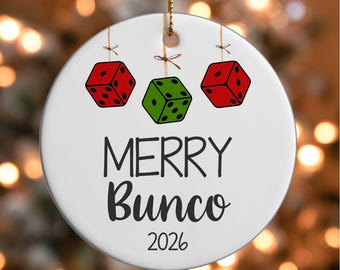 May include: A round white ceramic ornament featuring a graphic of two red dice and one green die hanging from strings. The text MERRY BUNCO is printed in black, followed by the year 2026 in a simple black font. A gold string is attached at the top.