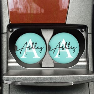Puede incluir: Dos posavasos turquesas para coche con el nombre "Ashley" en escritura cursiva y una letra "A" blanca. Los posavasos están en los portavasos de un coche, con un interior marrón y plateado.