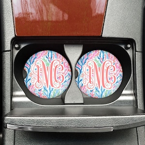 May include: Two round car coasters with a floral pattern in pink, blue, and green. Each coaster features a coral-colored monogram letter "M" in the center. The coasters are placed in a car's cup holders.