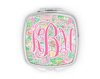 May include: A square silver compact mirror featuring a vibrant, multi-colored floral and architectural pattern on the lid. Centered on the lid is a large, stylized hot pink monogram with the letters KBM in an ornate, cursive script font.