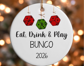 May include: A round white ceramic ornament featuring two red dice and one green die hanging from strings. The text reads Eat, Drink & Play BUNCO and the year 2026. The ornament is suspended by a gold cord at the top for hanging on a tree.