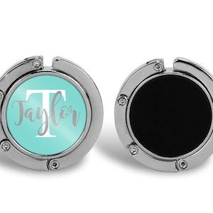 May include: A silver-colored, round, foldable handbag hook with a turquoise blue background and the name "Taylor" in white lettering with a white capital "T". The hook is closed in the image.