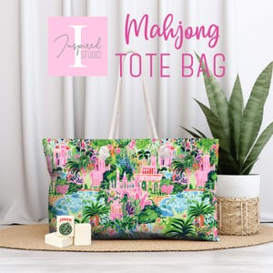 May include: A rectangular tote bag featuring a vibrant, multi-colored tropical print with pink buildings, green palm trees, and lush foliage. The bag has two white shoulder straps. A white Mahjong tile with a green Joker design sits in front.