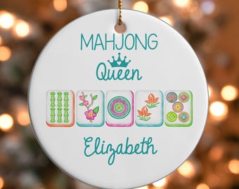 May include: A round white ceramic ornament featuring the teal text MAHJONG Queen and the name Elizabeth. Below the text are five rectangular mahjong tiles displaying green bamboo, a pink flower branch, a circular mandala, orange flowers, and colorful circles.