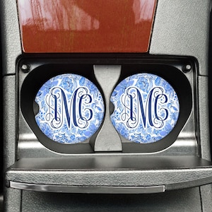 May include: Two round car coasters with a blue floral pattern and a navy blue monogram "MC" are placed in a car's cup holders. The coasters have a white background with blue floral designs.