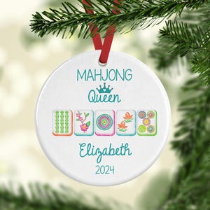 May include: White round Christmas bauble featuring a green, pink and blue Mahjong tile design. The bauble reads "Mahjong Queen Elizabeth 2024".