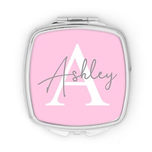 May include: A square, silver compact mirror with a pink background. The mirror features a large white letter "A" with the name "Ashley" in gray script. The compact is closed and ready for use.