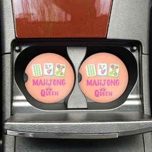 May include: Two peach-colored car coasters with the text "MAHJONG QUEEN" in pink and white. Each coaster features a Mahjong tile design. The coasters are in a car's cup holders.