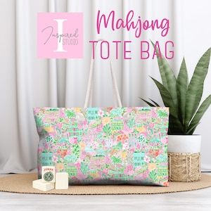 May include: A tote bag featuring a vibrant, multi-colored pattern of tropical foliage, flowers, and buildings in shades of pink, green, and yellow. The bag has two white shoulder straps and is positioned near a white Joker mahjong tile.