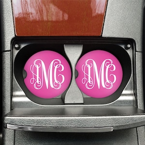 May include: Two pink car coasters with a white monogram letter "M" in a car's cup holder. The coasters are round and have a glossy finish. The car's interior features a black, silver, and brown color scheme.