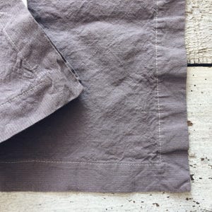 May include: A gray linen napkin with a white stitched border. The napkin is folded in half and the edges are visible.