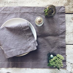 May include: A gray linen placemat with a white plate and a folded gray linen napkin. There are two small glass jars with lids on the placemat, one with green sprigs and the other with a silver lid.