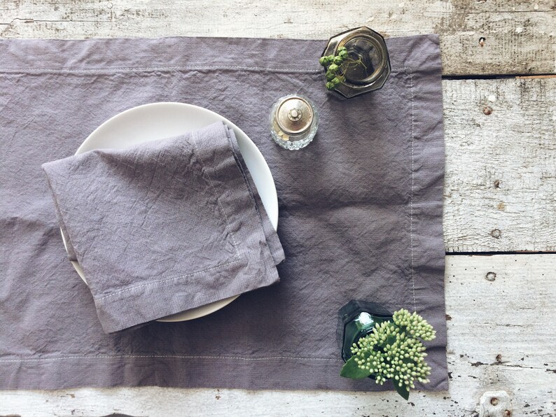 May include: A gray linen placemat with a white plate and a folded gray linen napkin. There are two small glass jars with lids on the placemat, one with green sprigs and the other with a silver lid.