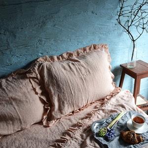 May include: A pink linen bedding set with ruffled edges, featuring a pillow and a duvet cover. A silver tray with a cup of tea, a pastry, and flowers sits on the bed.