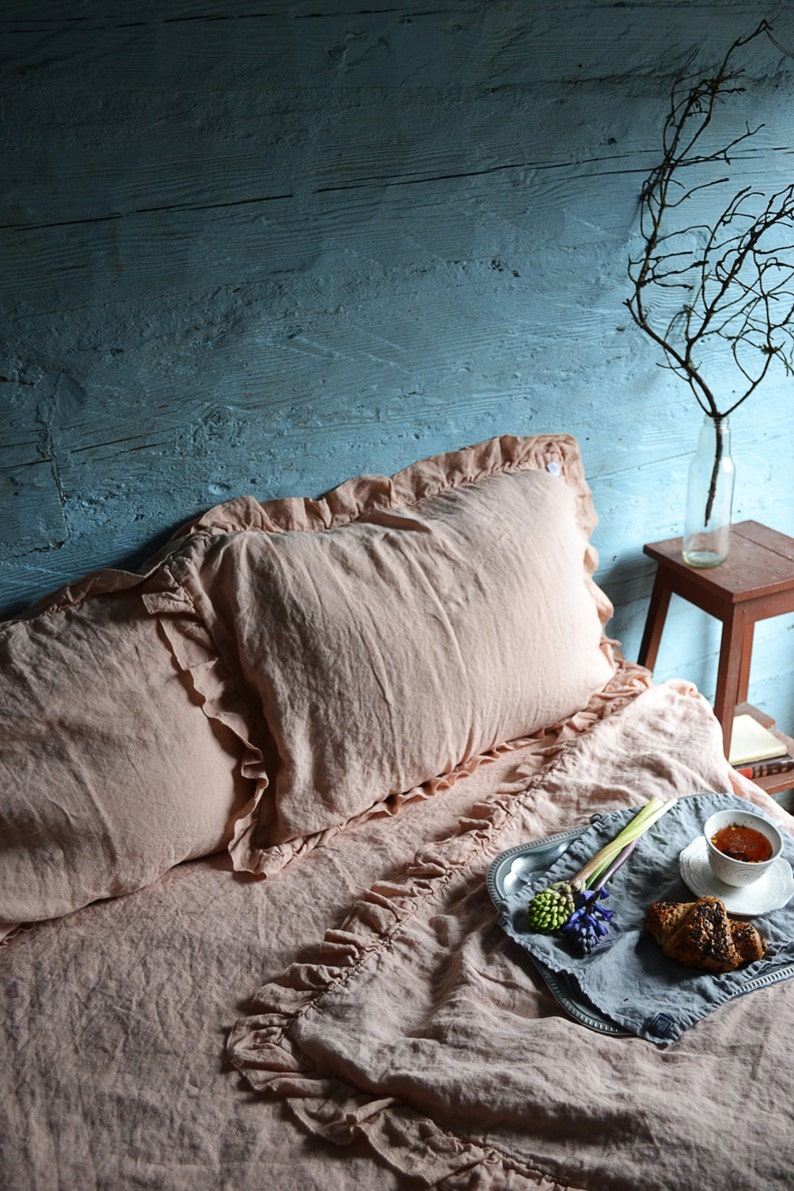May include: A pink linen bedding set with ruffled edges, featuring a pillow and a duvet cover. A silver tray with a cup of tea, a pastry, and flowers sits on the bed.