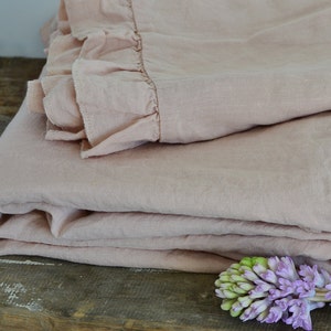 May include: A soft, blush pink linen throw blanket with a ruffled edge. The blanket is folded and draped over a rustic wooden surface with a purple flower in the foreground.