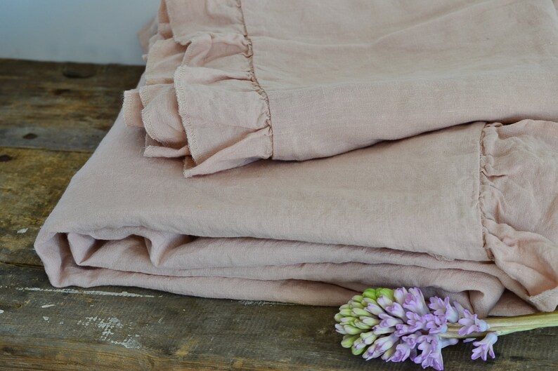 May include: A soft, blush pink linen throw blanket with a ruffled edge. The blanket is folded and draped over a rustic wooden surface with a purple flower in the foreground.