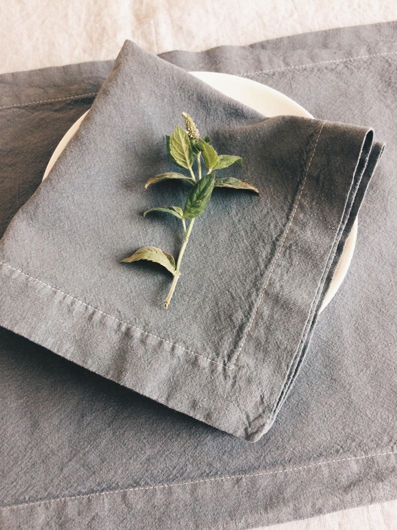 May include: A gray linen napkin folded on a gray linen placemat with a sprig of green foliage on top.