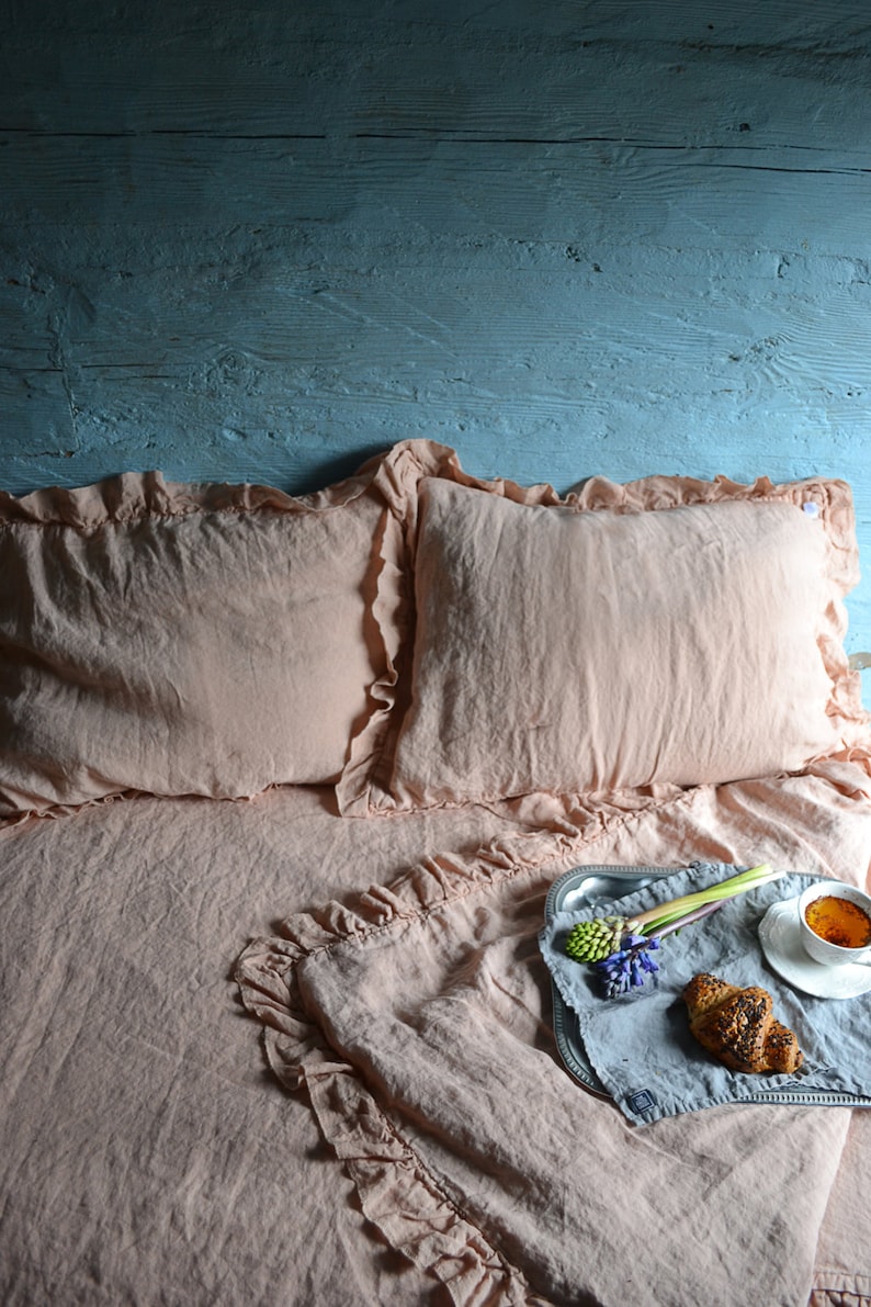 May include: A pink linen bedding set with ruffled edges. The bedding is arranged on a bed with a blue wooden headboard. A silver tray with a croissant, flowers, and a cup of tea is on the bed.