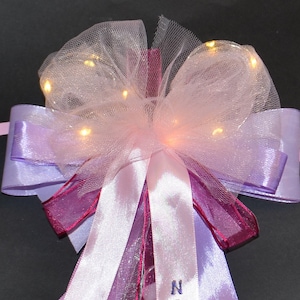 Op de afbeelding: Een roze en paarse strik met een doorschijnende overlay en witte lampjes. De strik heeft een satijnen lint met het woord "NAME" erop geborduurd.